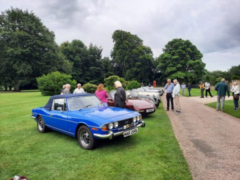 A blue Triumph Stag convertible parked on grass with people in the background.