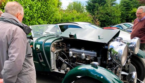 A green Lagonda LG45 Drophead Coupe with its hood open, revealing the engine, surrounded by people.
