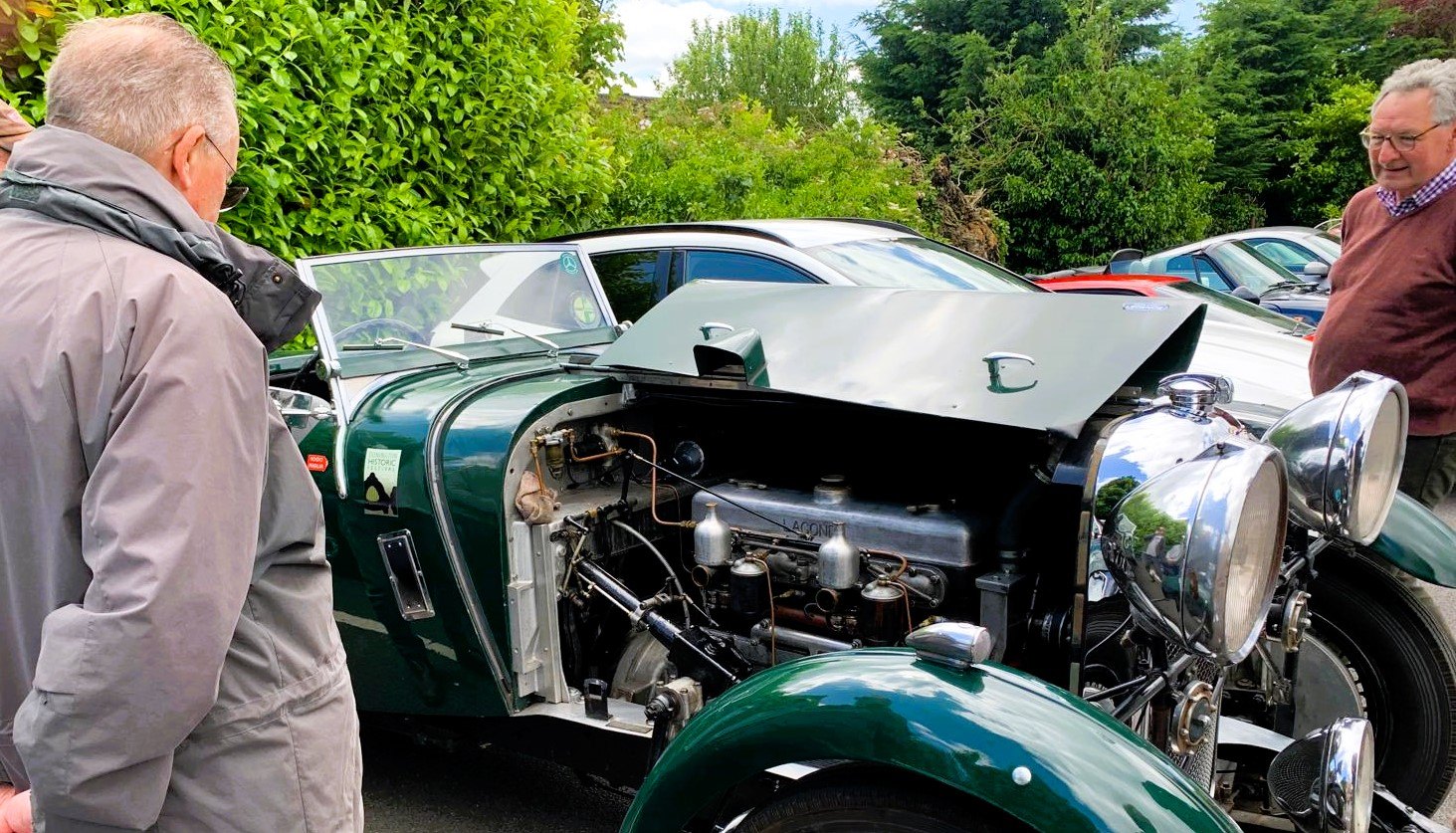 A green Lagonda LG45 Drophead Coupe with its hood open, revealing the engine, surrounded by people.