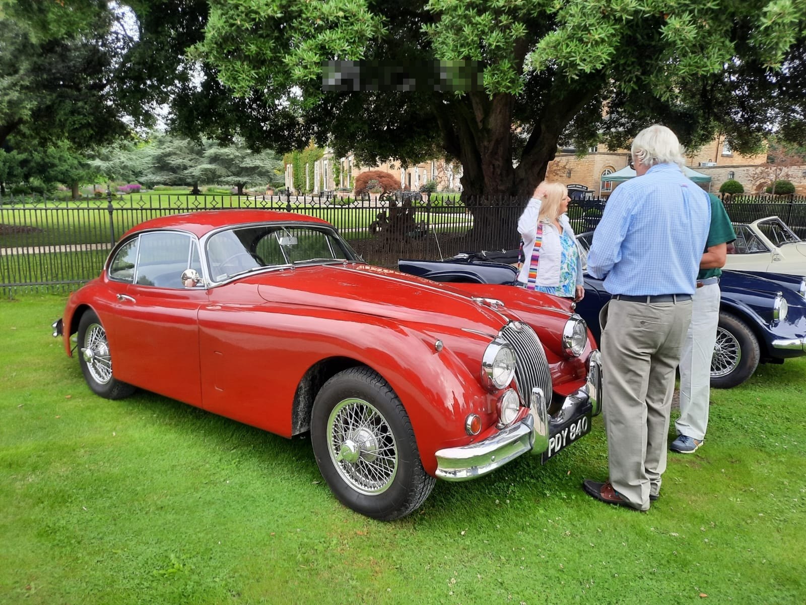 Red Jaguar XK140 Coupé parked in a green field with onlookers nearby.